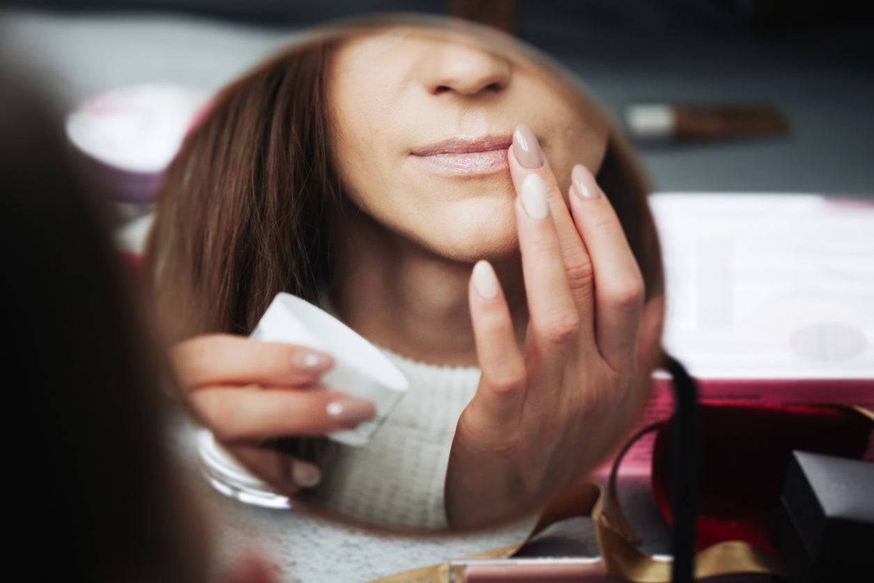 A woman putting petroleum gelly on her chapped lips in front of a mirror.